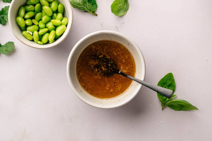 A white bowl of brown vinaigrette with a spoon sits beside a bowl of fresh green edamame on a light surface, garnished with scattered basil leaves.
